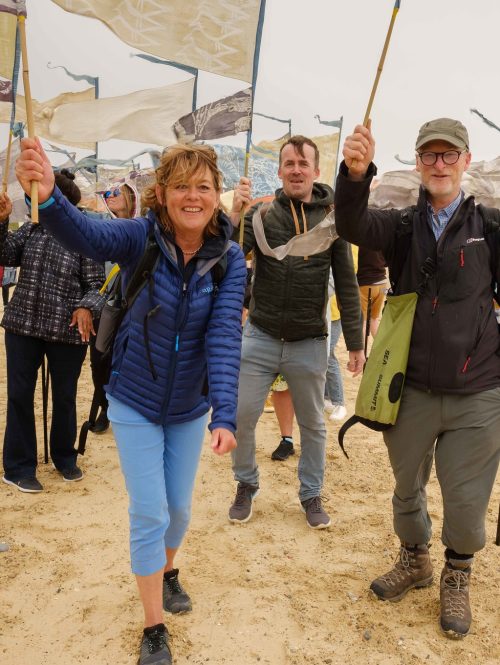 A group of people on a beach holding silk pennants in different colours.