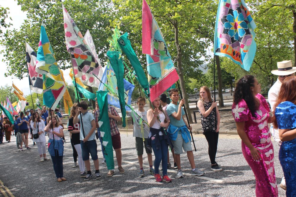 A group of young people are walking with a set of colourful flags held in the air as part of a carnival parade.