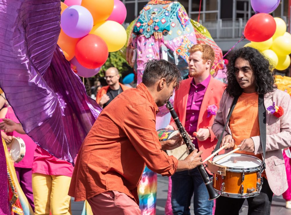A group of musicians in colourful clothes perform outdoors surrounded by balloons and flags.