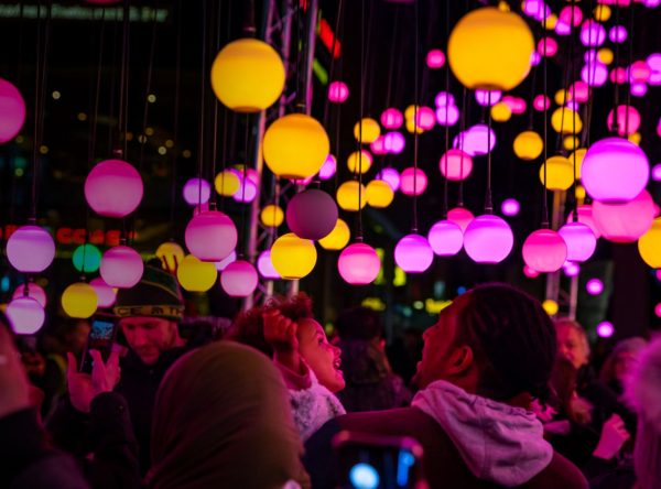 Audiences looking at a light installation by night, consisting of light bulbs in pink and orange hanging over the audiences's heads.