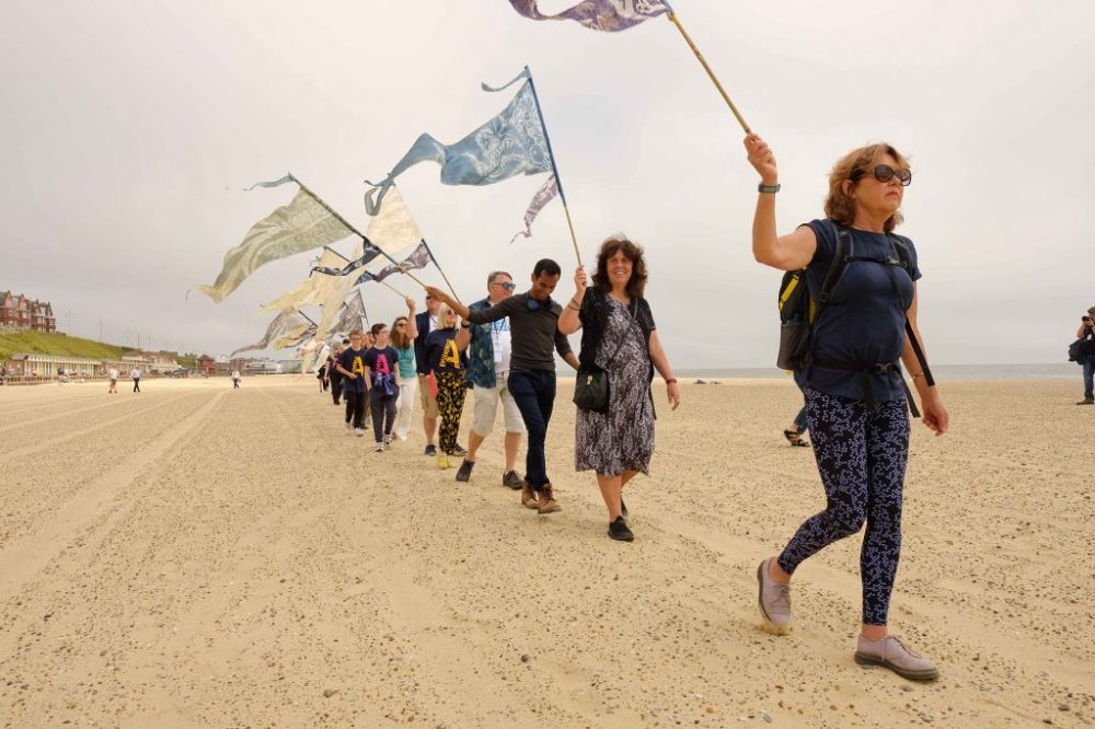 A group of people are walking along a beach holding colourful pennants in the air.