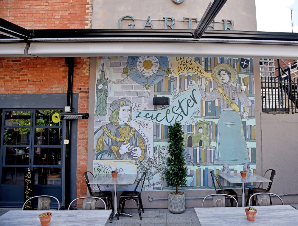 This image shows a huge outdoor mural by Rosie and Ramona at the Knight and Garter in Leicester. The Mural was hand illustrated and printed onto vinyl then fixed to the wall. The mural has the words Leicester, and, Deeds Not Words, written on it and features hand-drawn historical references to Leicester. The mural features King Richard III, Alice Hawkins and the Clock Tower.