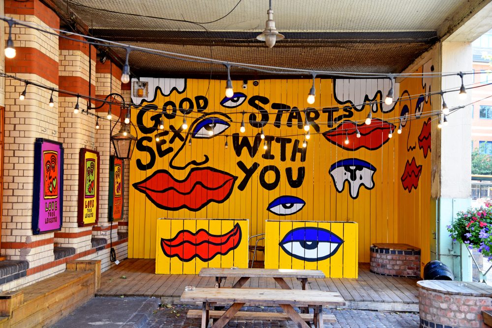 This image shows an outdoor painting by Katie Fishlock at The Parcel Yard in Leicester. The painting's background is bright yellow and covers a whole wooden-partition wall and some plant containers. The yellow background has eyes, lips and other facial elelements painted on it in an abstract fashion. The text that is painted reads, Good sex starts with you.
