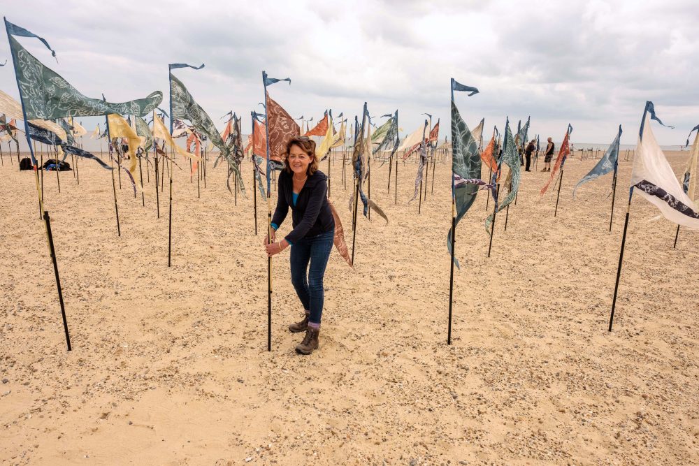 A person on a beach standing next to lots of silk pennants in different colours.