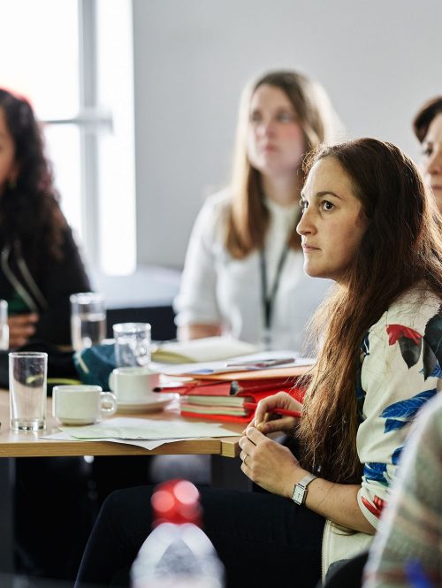 A group of women sit at a desk watching some give a presentation.