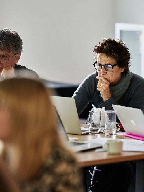 A woman focuses on her laptop in a meeting, there are other people in the meeting.