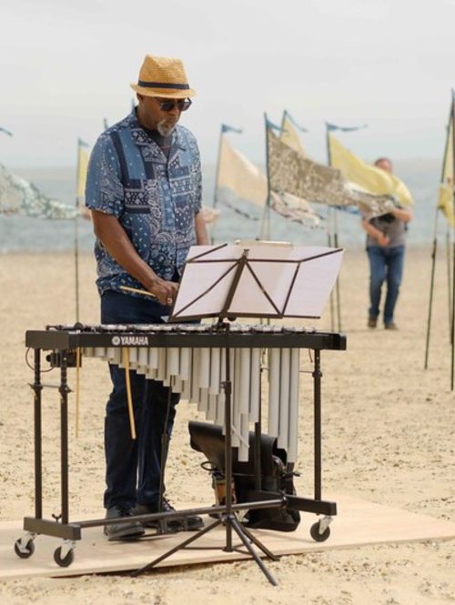 A man wearing a hat and sunglasses plays on a xylophone on a beach. In the background is a series of painted silk flags decorating the space.