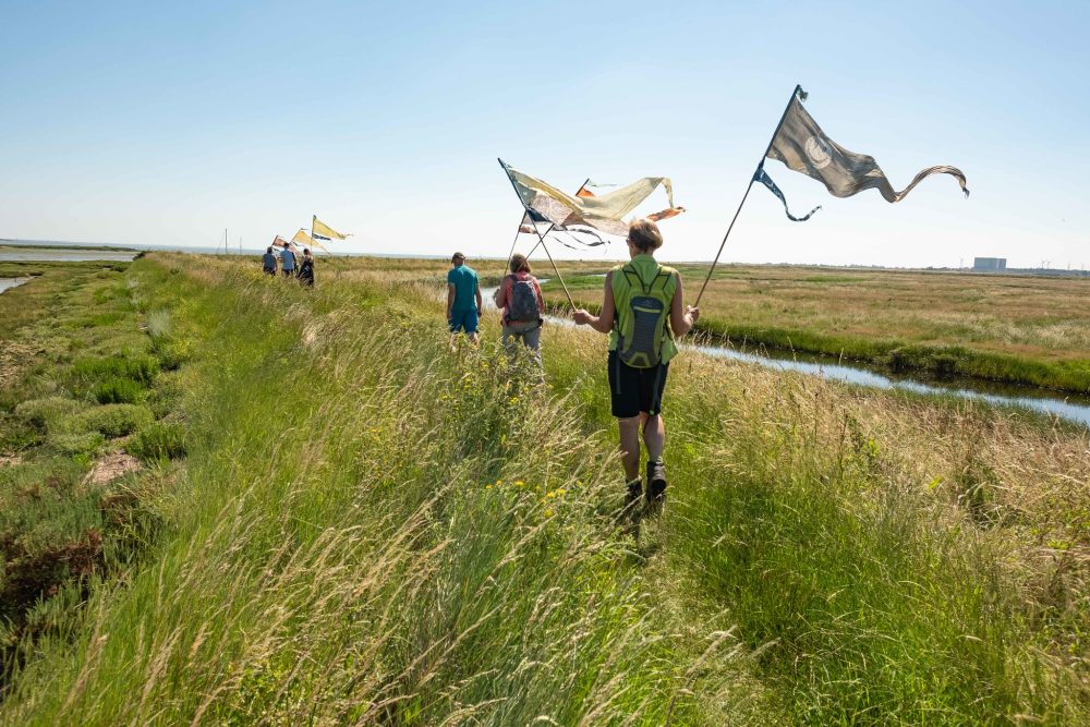 In a wide open space near a river people are walking with painted silk flags in their hand and the wind is blowing the flags in the air.