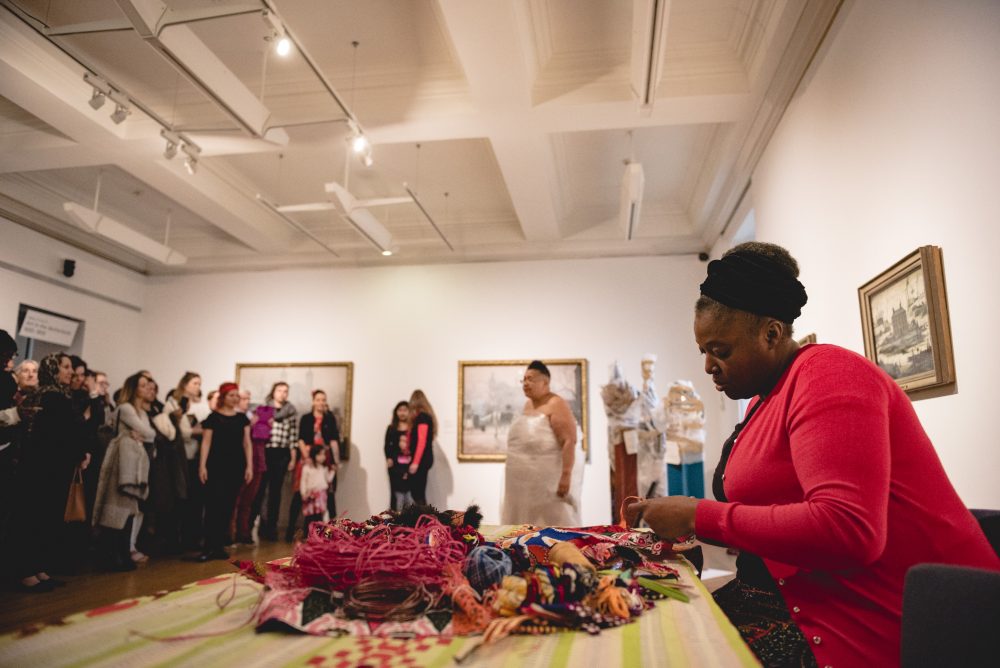 A performer working with fabric threads on a table in an indoor exhibition space.