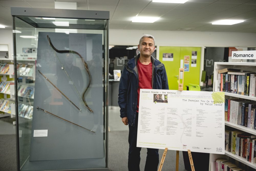 An artist stands next to an information sign and a glass cabinet with a bow and arrow in a library.