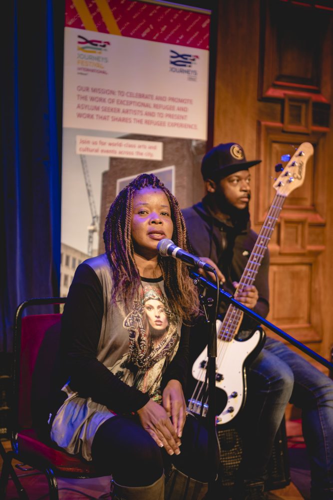 This image shows a woman singing into a microphone and behind is a man with a baseball cap playing bass guitar. They are standing in front of a banner for Journeys Festival International.