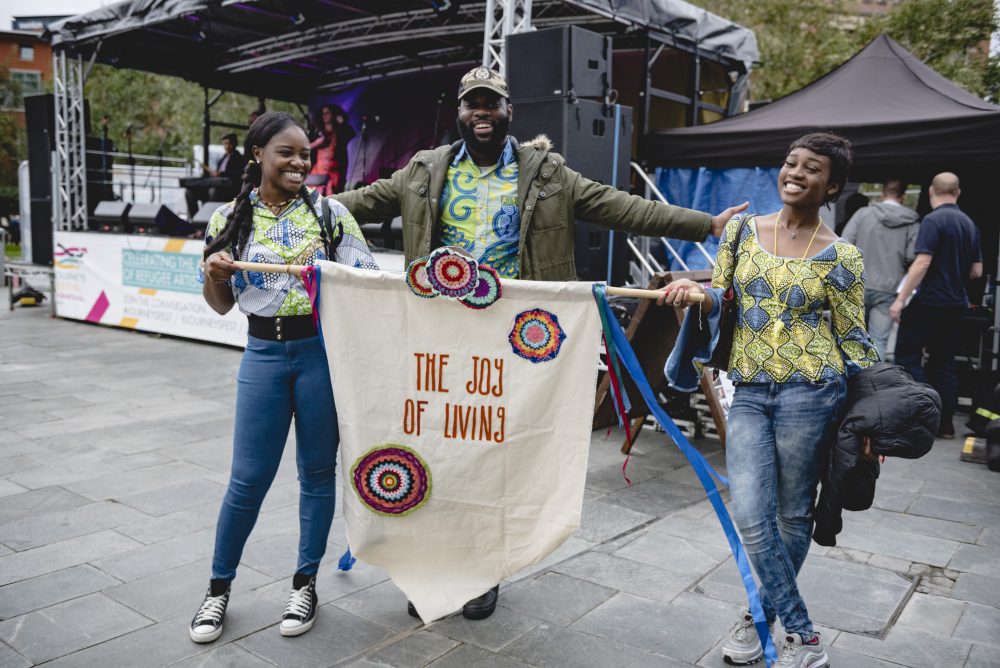 Three people in front of a festival music stage holding a banner saying The Joy of Living.