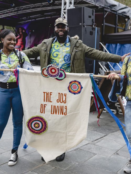 Three people in front of a festival music stage holding a banner saying The Joy of Living.