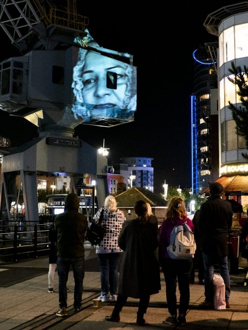 This image shows the portrait of a woman wearing a head scarf that is being projected onto a crane at Gun Wharf in Portsmouth.