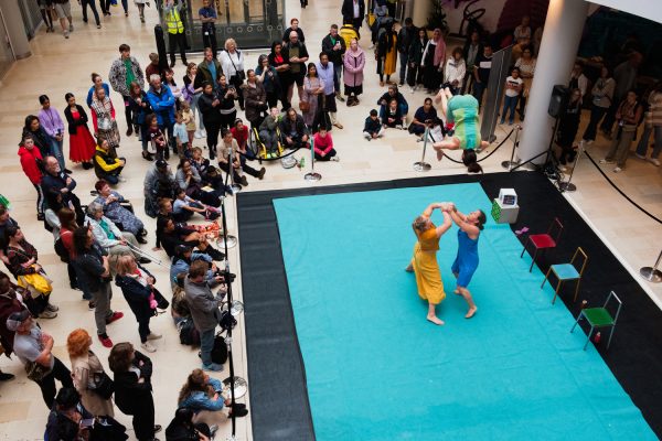 Two performers throwing another performer into the air on an indoor dance stage, surrounded by audiences.
