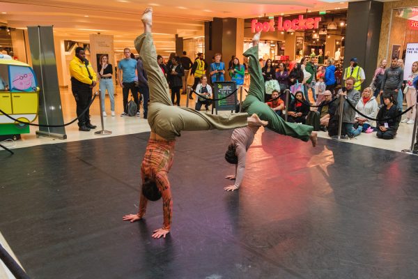 Two performers in a handstand dance pose in an indoor space, surrounded by audiences.