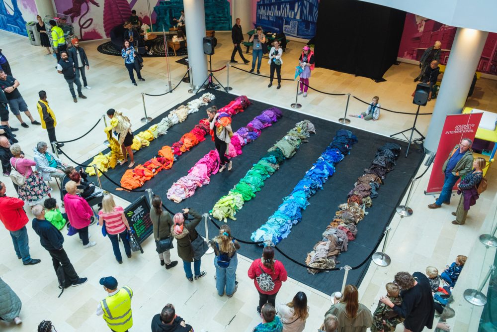 Bird's view perspective of two performers on an indoor square between lanes of differently coloured clothes, surrounded by audience members.