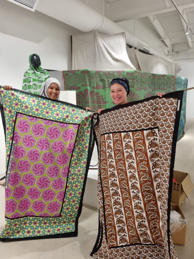 Two women are holding up highliy detailed and colourful peices of fabric in an exhibition space.