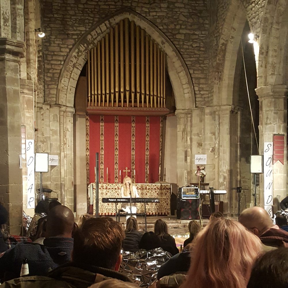 Audiences in a church listening to a concert by a performer singing and playing on a keyboard.
