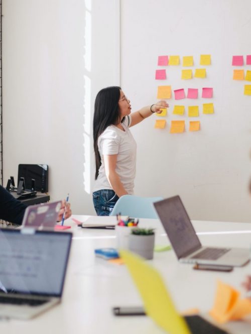 A woman in a meeting pointing to a white board with post-it notes on it.