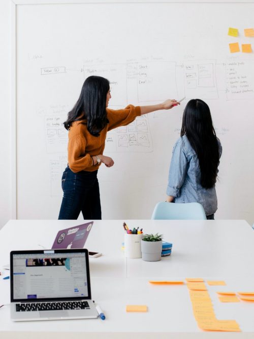 Two woman are standing at a white board in a meeting, the white board has post it notes on.