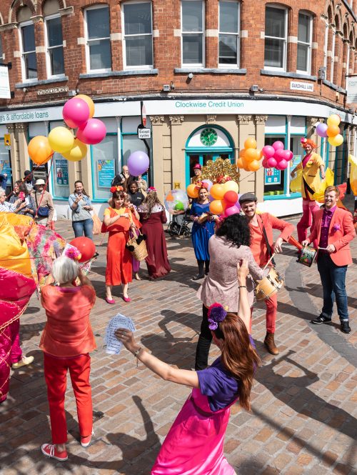 A group of performers in colourful costumes and balloons dancing in an outdoor square.