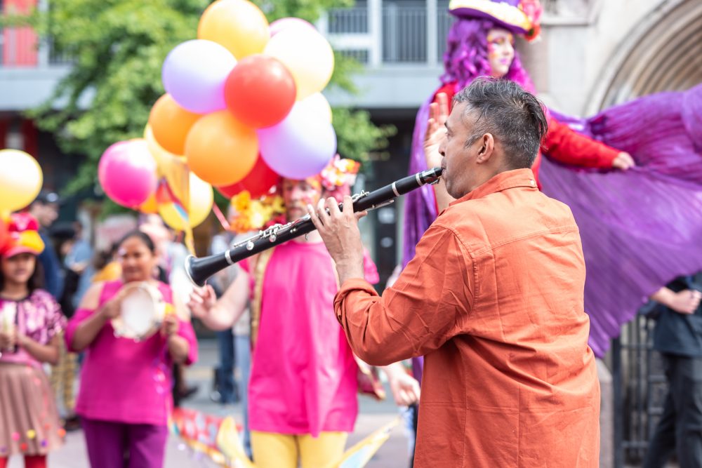 A musician in an orange suit is playing a clarinet at an outdoor parade. In the background is a set of balloons and other people in the parade.
