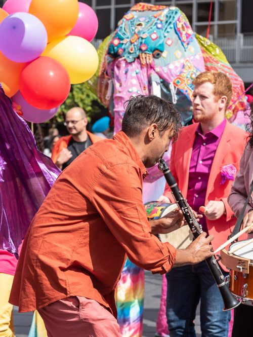 A group of musicians in very bright colourful clothing are playing in a carnival procession.