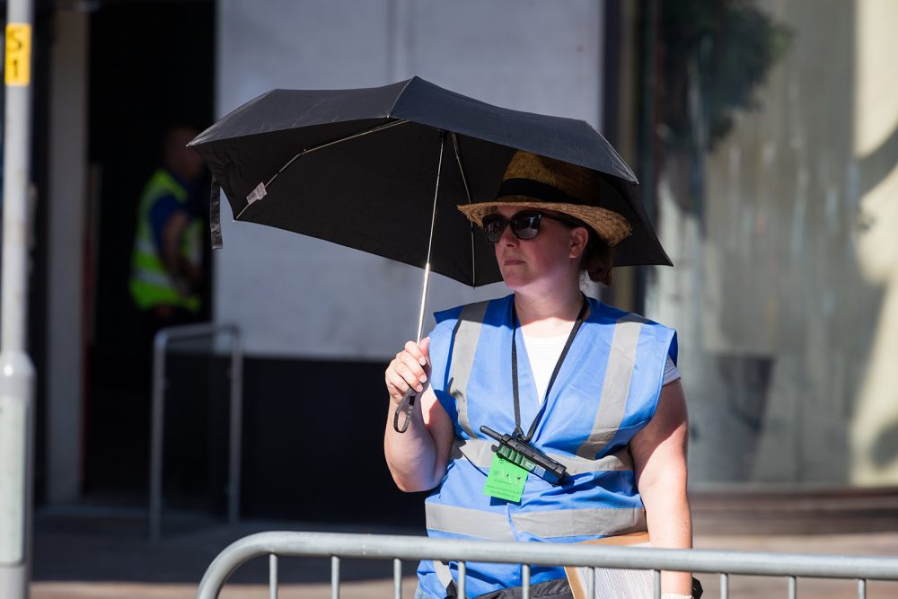 A person in a blue high-vis vest stands next to a barrier holding an umbrella in the sun.