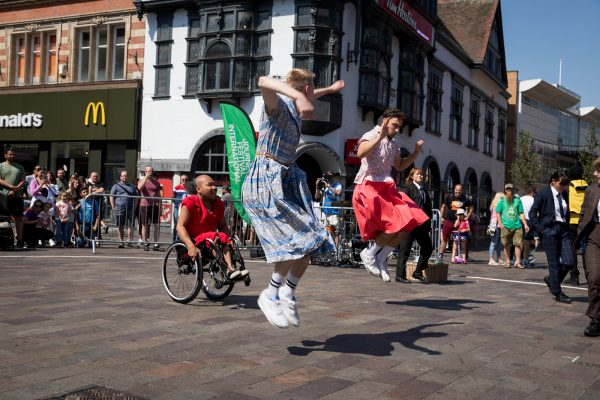Two dancers jumping into the air on an outdoor square, accompanied by a dancer in a wheelchair.