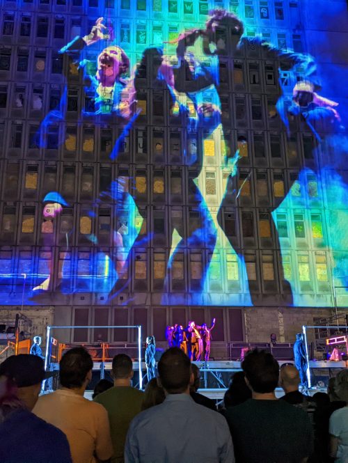 A crowd are watching projections against an old building. The projections are above an outdoor performance on a large stage.