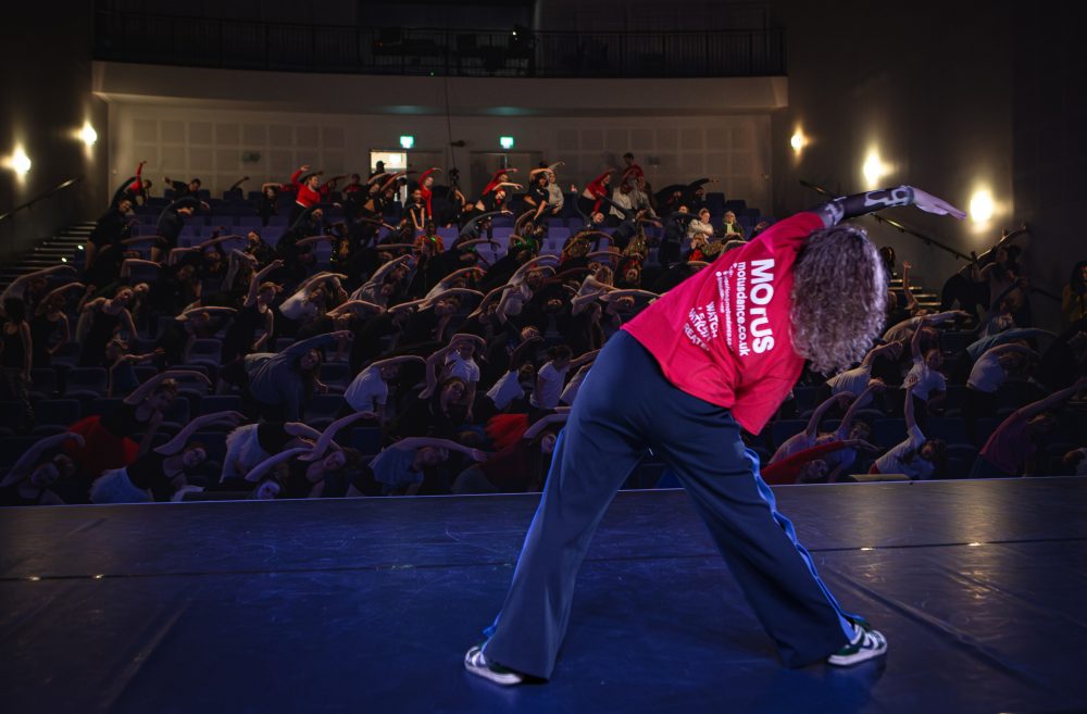 This image shows a dancer on stage facing an audience. The dancer is leading the audience through a seated warm-up exercise.