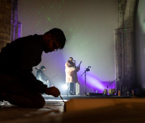 An artist painting on the floor, with a performer at a microphone in the background.