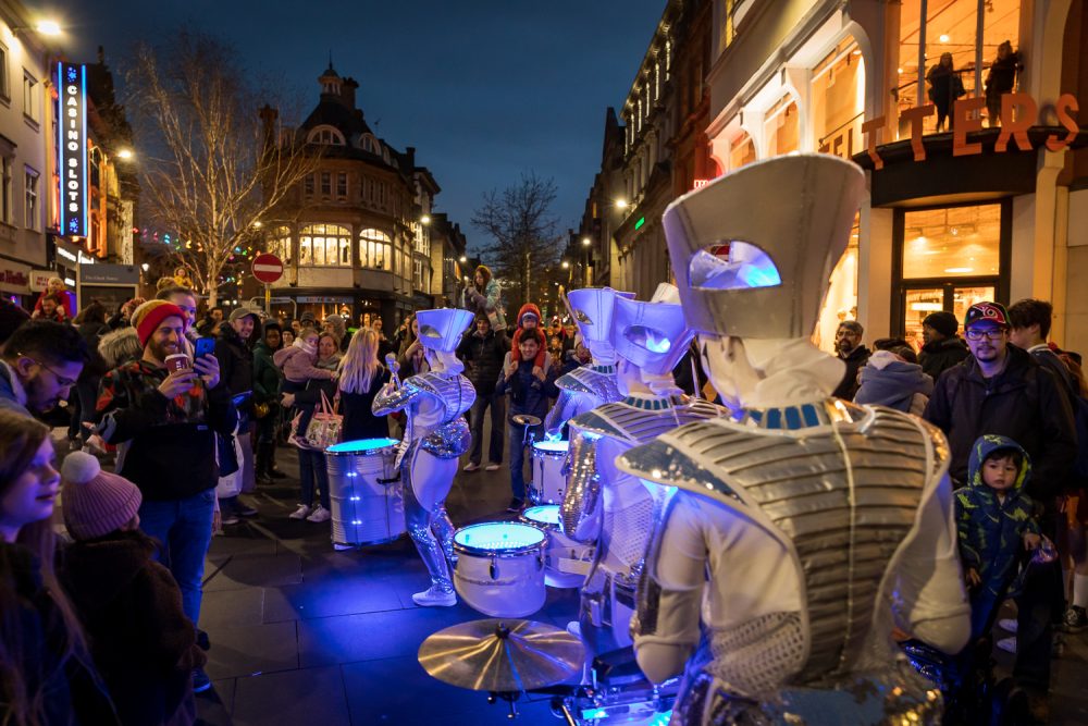 A group of drummers in costumes on an outdoor square by night, surrounded by audience members.