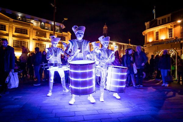 A group of men in costume are playing illuminated drums in a busy square.