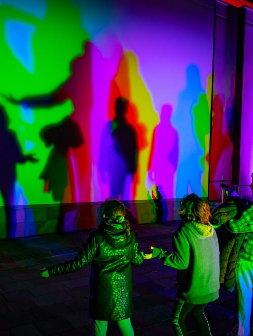 Audiences dancing in a light installation depicting their shadows in different colours on a wall.