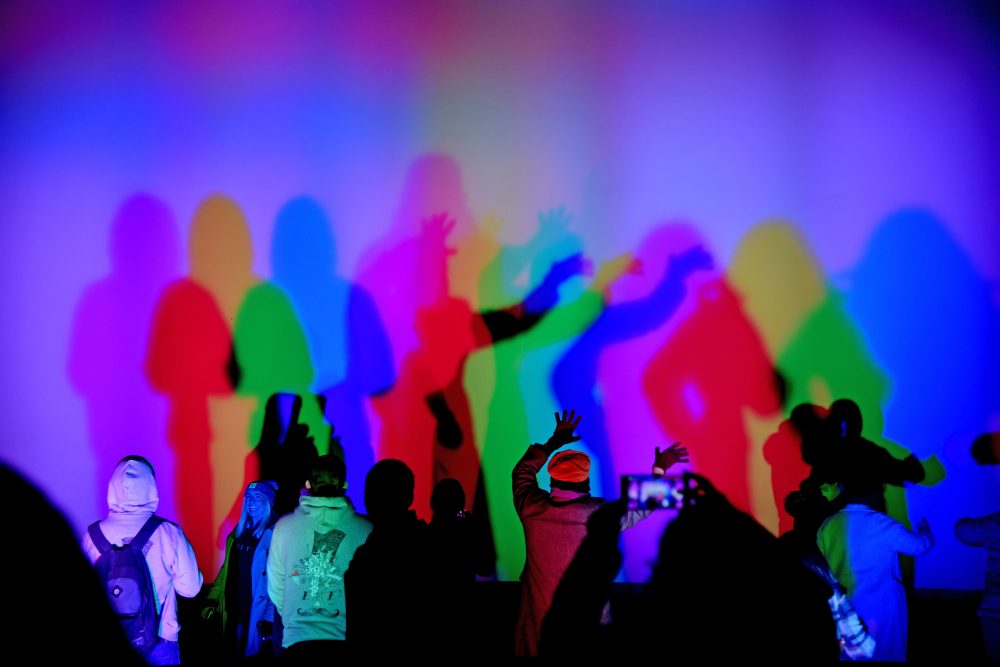 Audience members making shadow puppets on a colourfully lit outdoor wall.