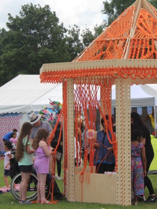 A tent with artistic knitted orange wool hanging down between the wooden frames.