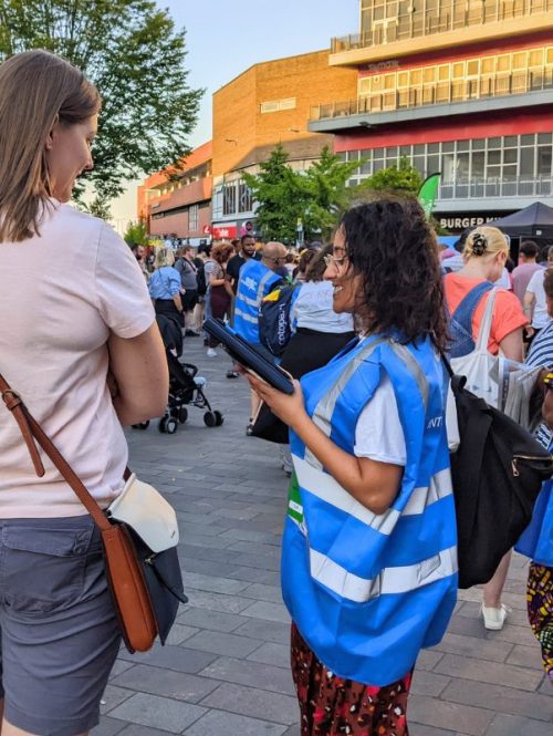 People in blue high-vis vests talk to audience members on an outdoor square.