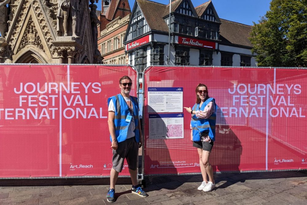 Two people in blue high-vis vests stand next to big red Journeys Festival International banners on an outdoor square.