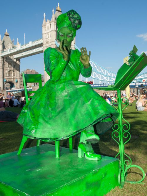 A living statue of a woman, her skin and clothes and hair are all bright green. She is sitting, posed, at a typewriter in front of Llondon Bridge.