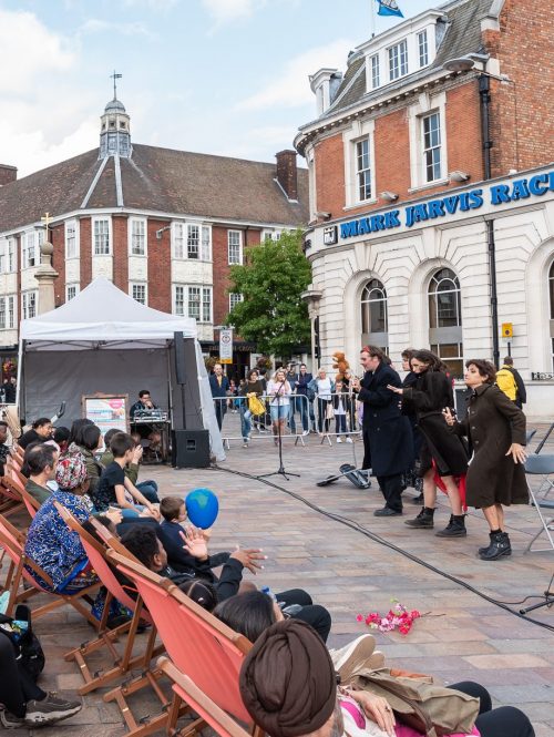 Performers on an outdoor square with audiences watching while sitting in canvas chairs