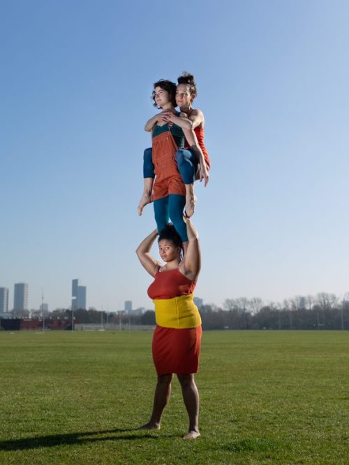 Three performers carrying each other in an acrobatic act in a park.