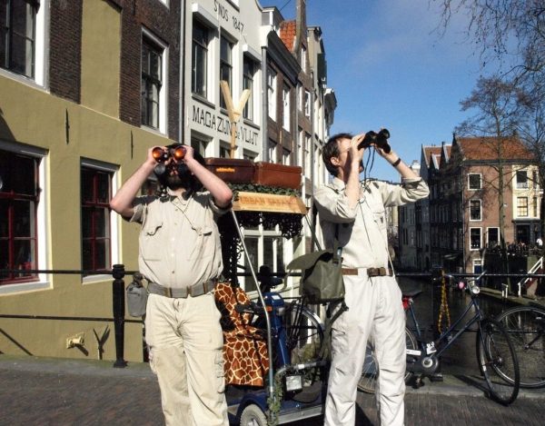 Two performers in beige safari outfits look at an urban street with their binoculars.