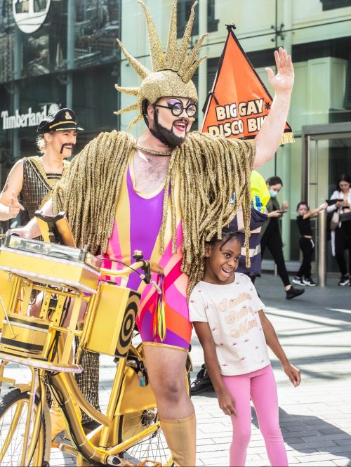 Performers in elaborate costumes on a shopping street, with one of them on a golden bike posing for a picture with a child.