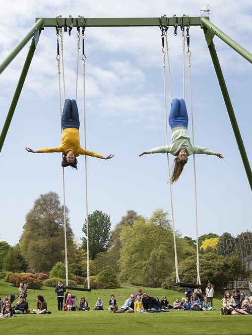 Two performers hanging upside down on a massive swing in a park.