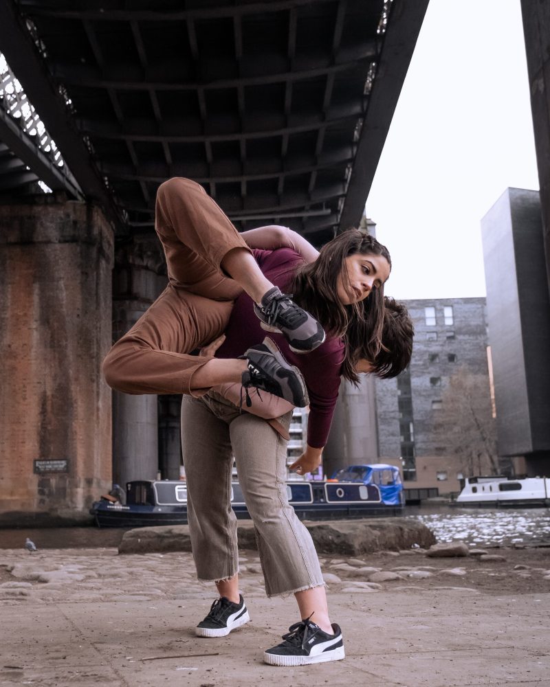 Two performers in a dance position, where one carries the other, in an industrial area.