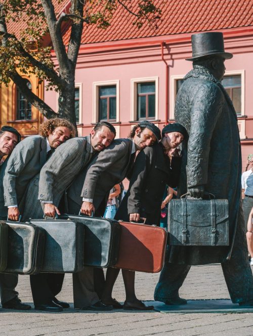 Eight performers with suitcases leaning on each others back towards a statue of a traveller.