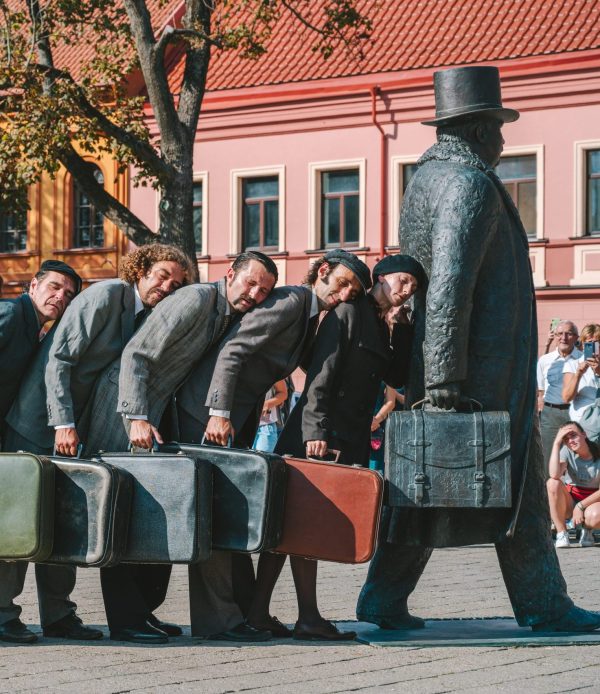 Eight performers with suitcases leaning on each others back towards a statue of a traveller.