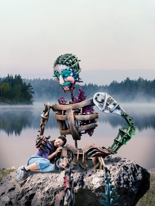 A giant puppet sitting on a rock on the shore of a lake, with a performer resting on its side.